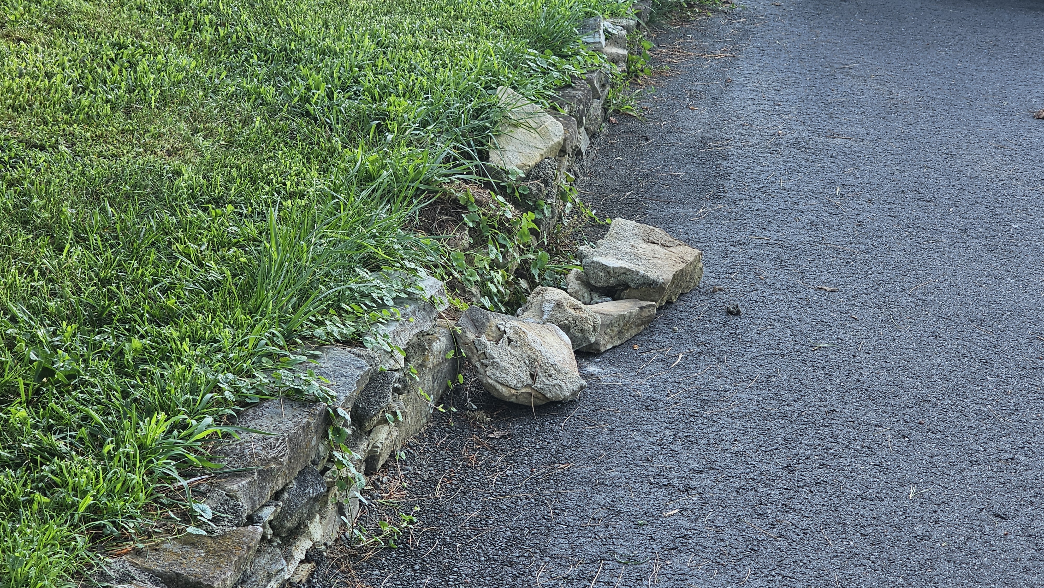 Small stone retaining wall with fallen rocks beside an asphalt driveway.